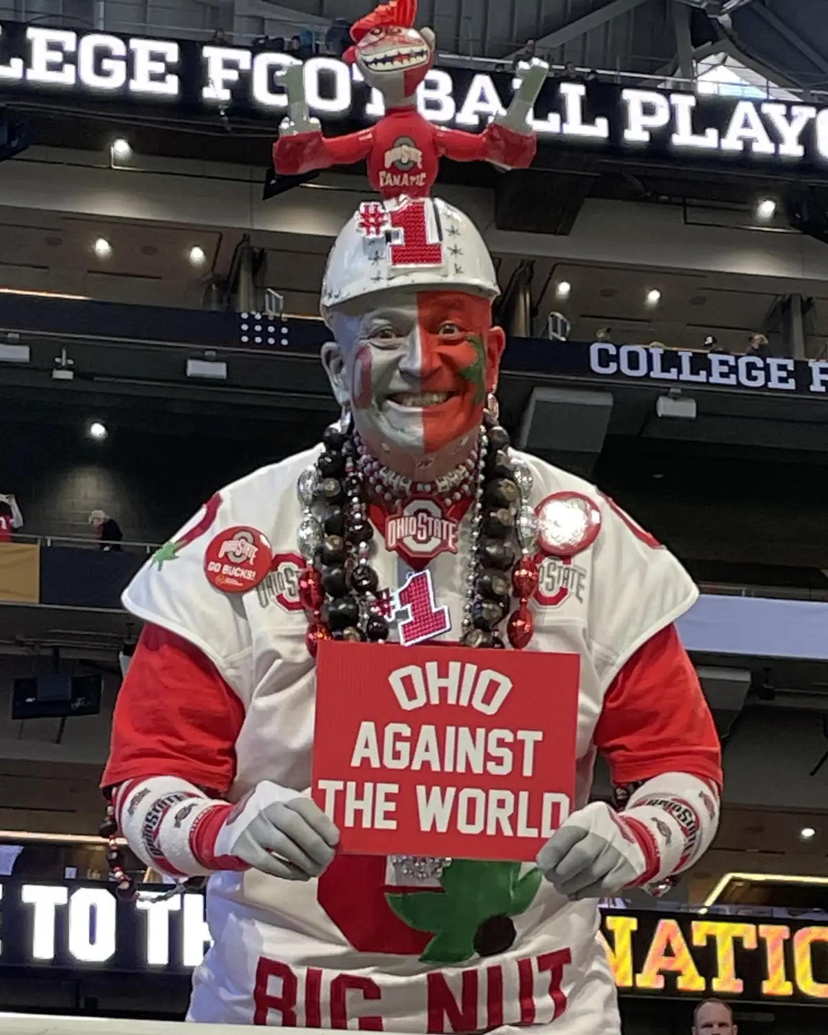 An Ohio State fan in half-scarlet face paint and a decorated hard hat holding a sign reading Ohio Against The World