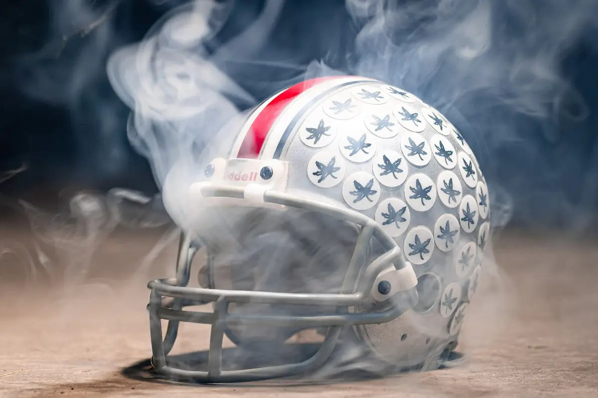 An Ohio State football helmet covered in buckeye leaf stickers, lit in drifting smoke against a dark background