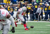 Ohio State tight end Cade Stover, number 8 in road white, bent toward a loose football on the turf with a Michigan defender beside him and the Michigan sideline behind