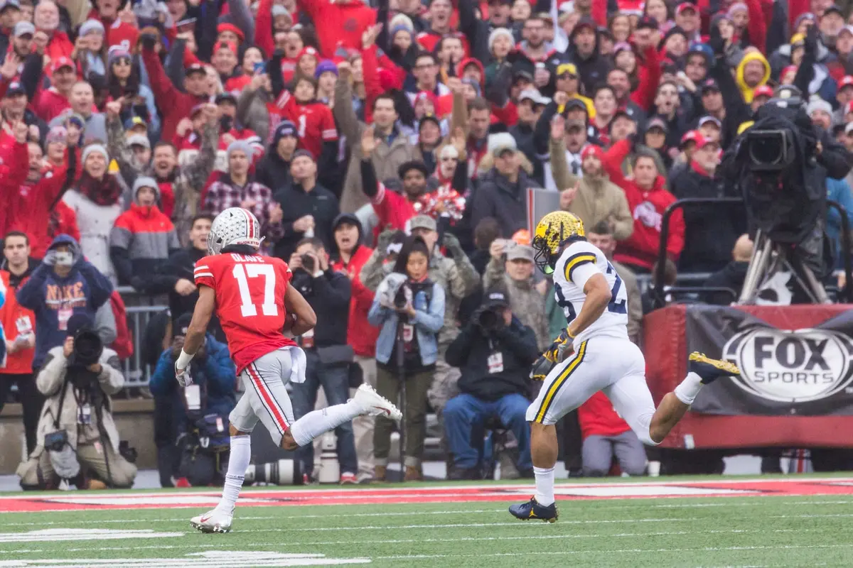 Ohio State receiver Chris Olave running upfield with the ball in a scarlet jersey, a Michigan defender trailing him and the crowd on its feet behind