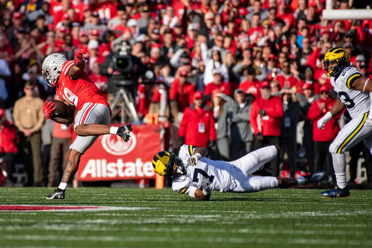 Ohio State running back DeaMonte Trayanum, number 19 in scarlet, cutting upfield with the ball tucked as a Michigan defender dives at his feet in front of a scarlet crowd