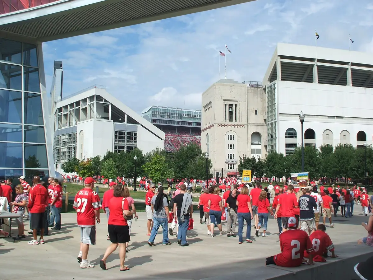 Fans in scarlet shirts and jerseys walking on a sunny plaza toward Ohio Stadium, the rotunda tower and open stands visible ahead