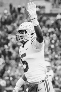 A black and white frame of an Ohio State receiver in road white, buckeye leaf stickers across his helmet, raising his right hand above his head