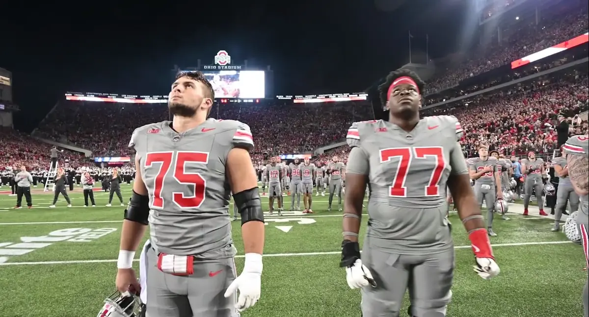 Two Ohio State offensive linemen in grey uniforms, numbers 75 and 77, standing on the field at a lit Ohio Stadium