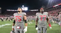 Two Ohio State offensive linemen in grey uniforms, numbers 75 and 77, standing on the field at a lit Ohio Stadium