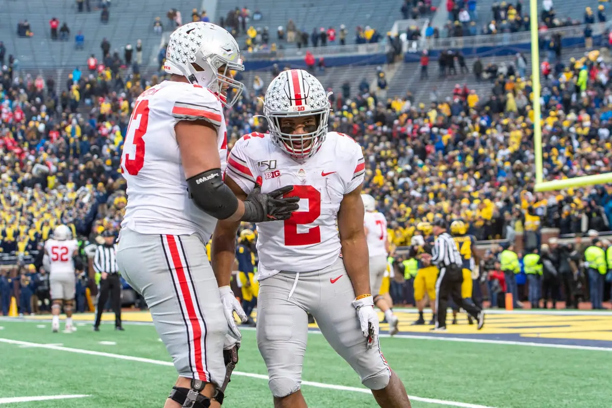 Two Ohio State players in white road jerseys celebrating together in the end zone, a maize and blue crowd behind them