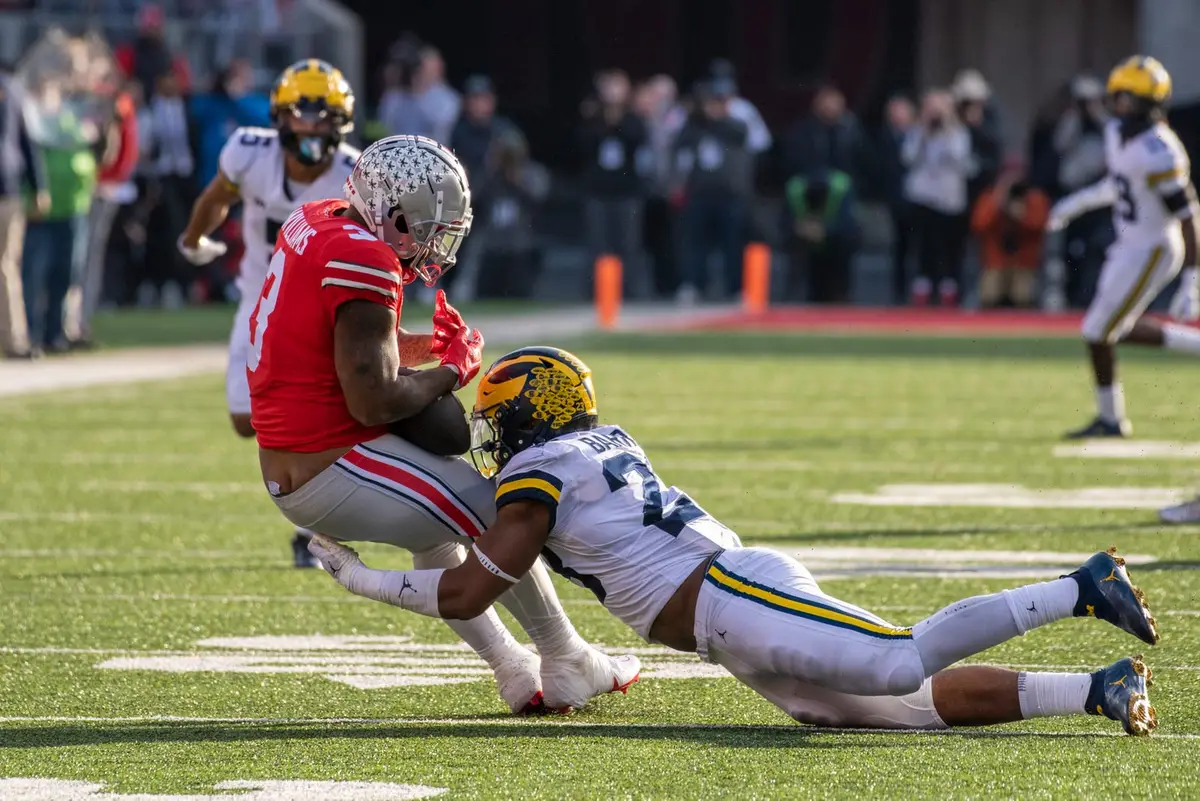 Ohio State running back Miyan Williams carrying the ball in scarlet as a Michigan defender wraps him up around the legs