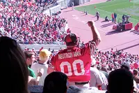 A fan in a scarlet Ohio State jersey reading NEUTRON MAN with the number 00 standing with one fist raised in the stands at Ohio Stadium