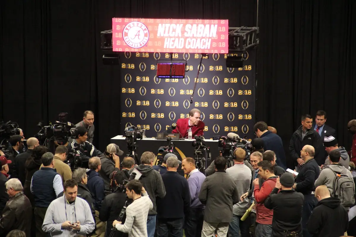 Nick Saban seated at a podium under an Alabama sign reading Nick Saban Head Coach, surrounded by a crowd of reporters and television cameras