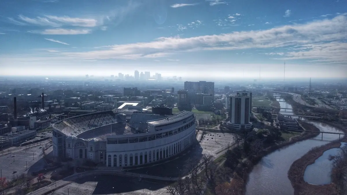 Aerial view of Ohio Stadium's curved exterior beside the Olentangy River, with campus buildings behind it and the downtown Columbus skyline in haze on the horizon under a blue sky
