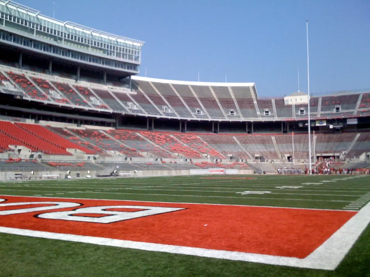 Ohio Stadium empty on a clear day, seen from field level at the corner of a scarlet end zone, the curved stands and a goalpost behind