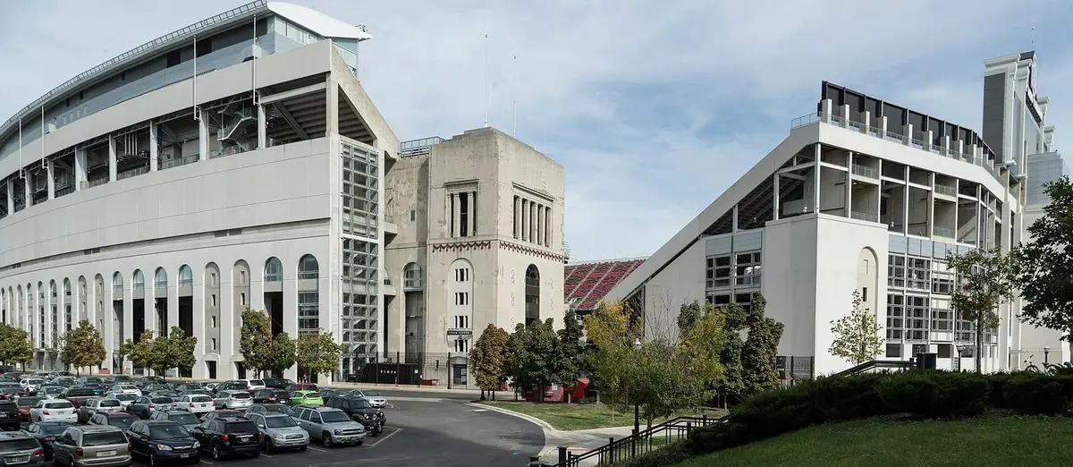The exterior of Ohio Stadium in Columbus, the arched windows of the original concrete stands on one side and a newer deck on the other, with scarlet seating visible through the gap between them