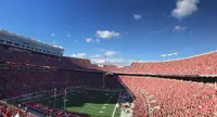 A packed Ohio Stadium in scarlet under a blue sky, seen from high in the stands, the horseshoe curving around the field toward the open end