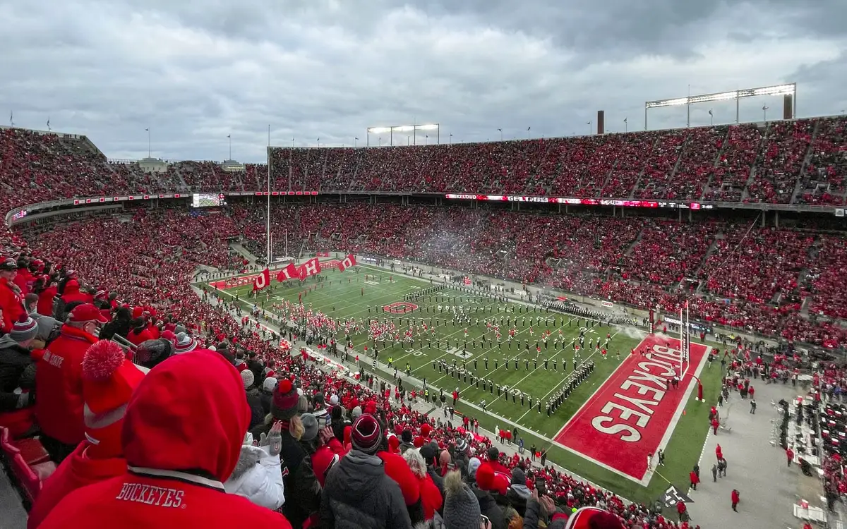 Ohio Stadium seen from the upper deck under a grey sky, the stands filled with fans in scarlet and the marching band spread across the field