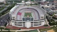 Aerial view into an empty Ohio Stadium over its south stands, the Ohio State logo and an OHIO STADIUM sign on top, the red and grey seats of the horseshoe wrapping the field beyond