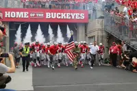 Ohio State players running out of the Ohio Stadium tunnel through jets of white smoke, under a red sign reading Home of the Buckeyes, fans crowding the rails above