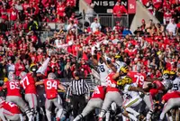 The Ohio State offensive line in scarlet holding a wall at the line of scrimmage while Michigan players leap with their arms up to block a kick, the ball in the air above them
