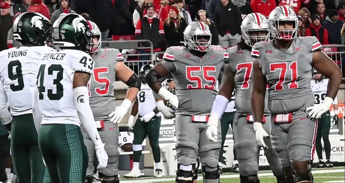 Ohio State linemen in grey uniforms standing near the line of scrimmage across from Michigan State players in white and green at Ohio Stadium