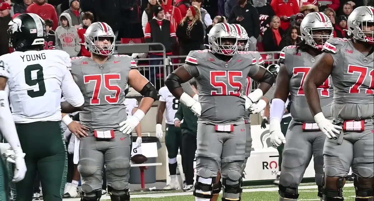 Ohio State offensive linemen in grey uniforms on the field with a Michigan State player at left