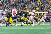 An Ohio State quarterback in a white road jersey pulls the ball back to throw as Michigan defenders in blue rush and a Buckeye lineman blocks