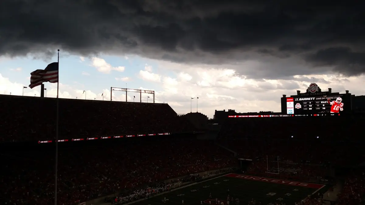 A dark storm front over a full Ohio Stadium, with the American flag flying and the scoreboard lit against the black sky