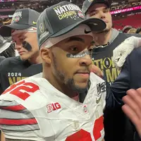 Ohio State running back TreVeyon Henderson in a National Champs cap and a scuffed white jersey on the field, teammates in championship shirts behind him